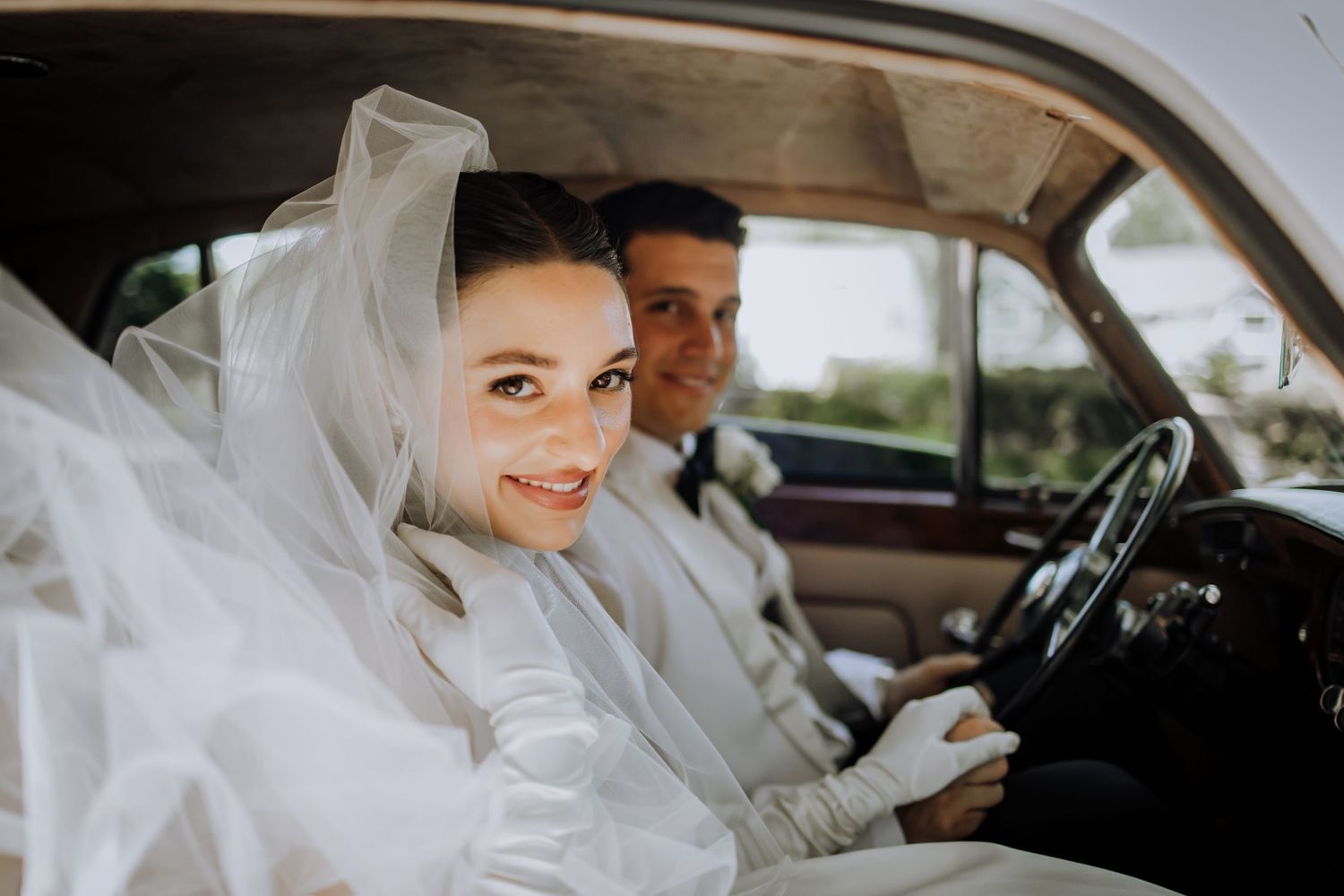 Bride in white veil and groom in suit sitting in a vintage car on their wedding day.