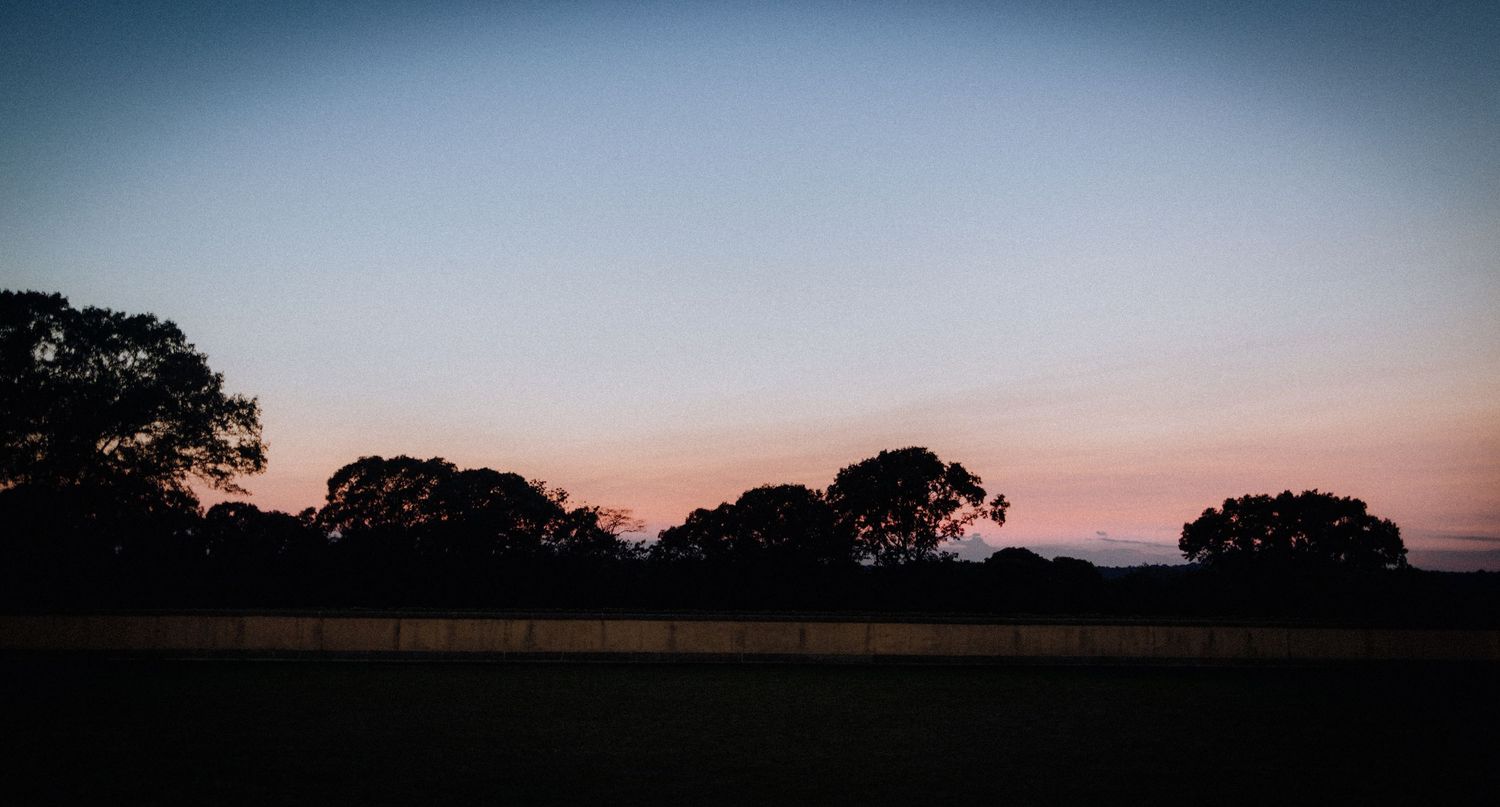 Silhouetted trees against a pink and blue dusk sky over a rural fence line at sunset.