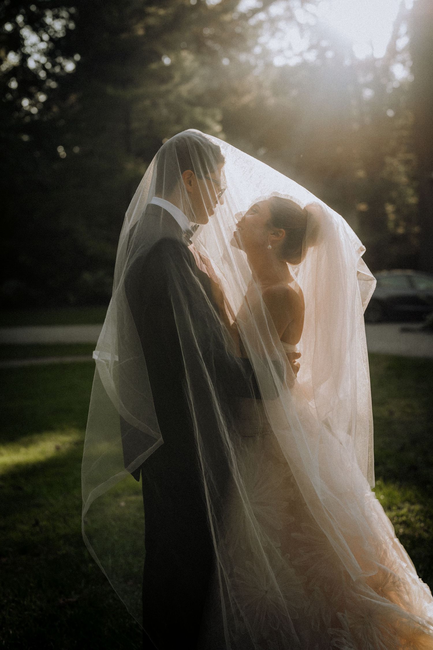 Bride and groom sharing a romantic moment under a flowing veil with golden sunlight glowing behind them outdoors.