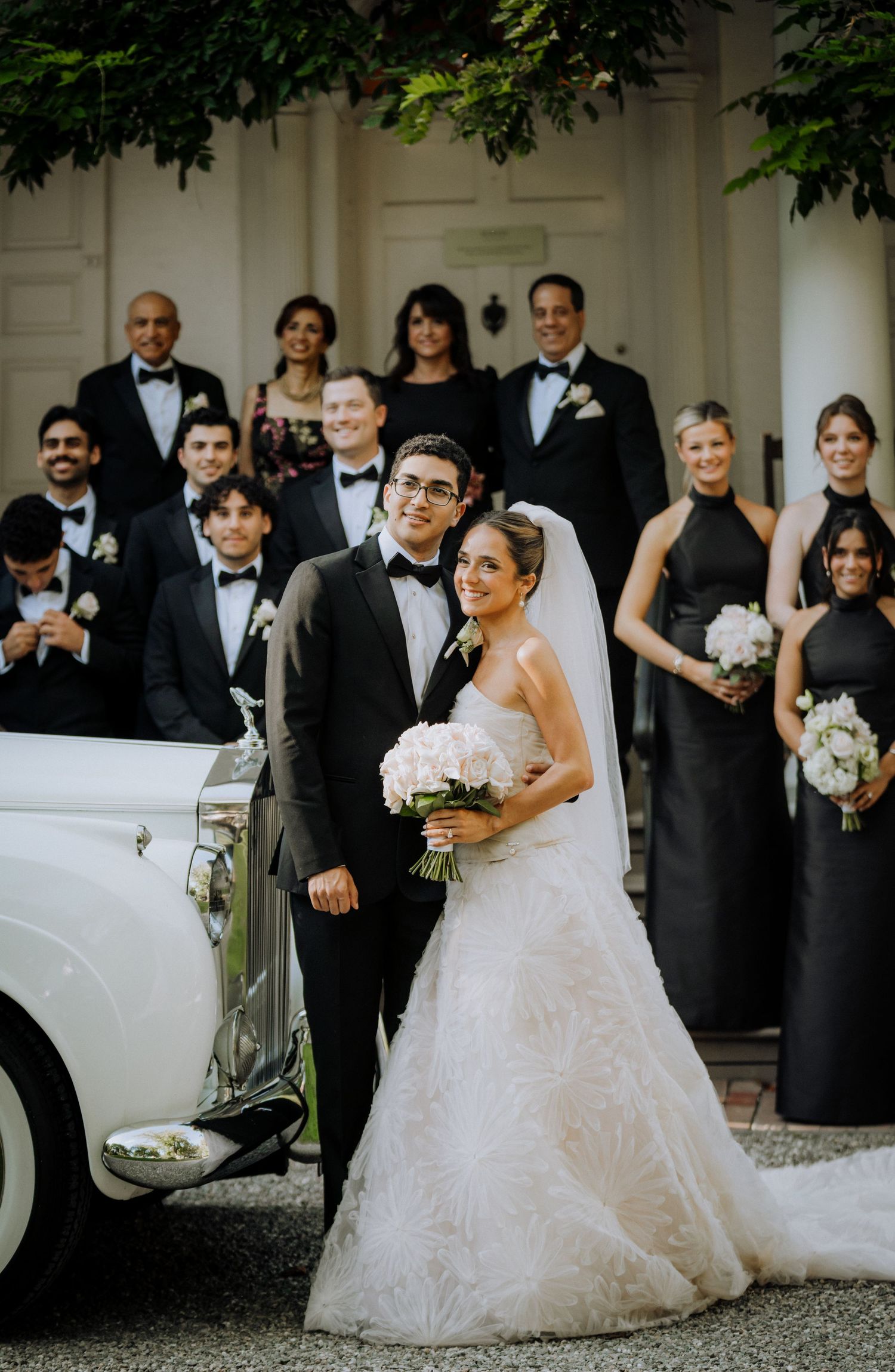 Bride and groom pose with wedding party in black attire beside a vintage white car at an elegant venue.