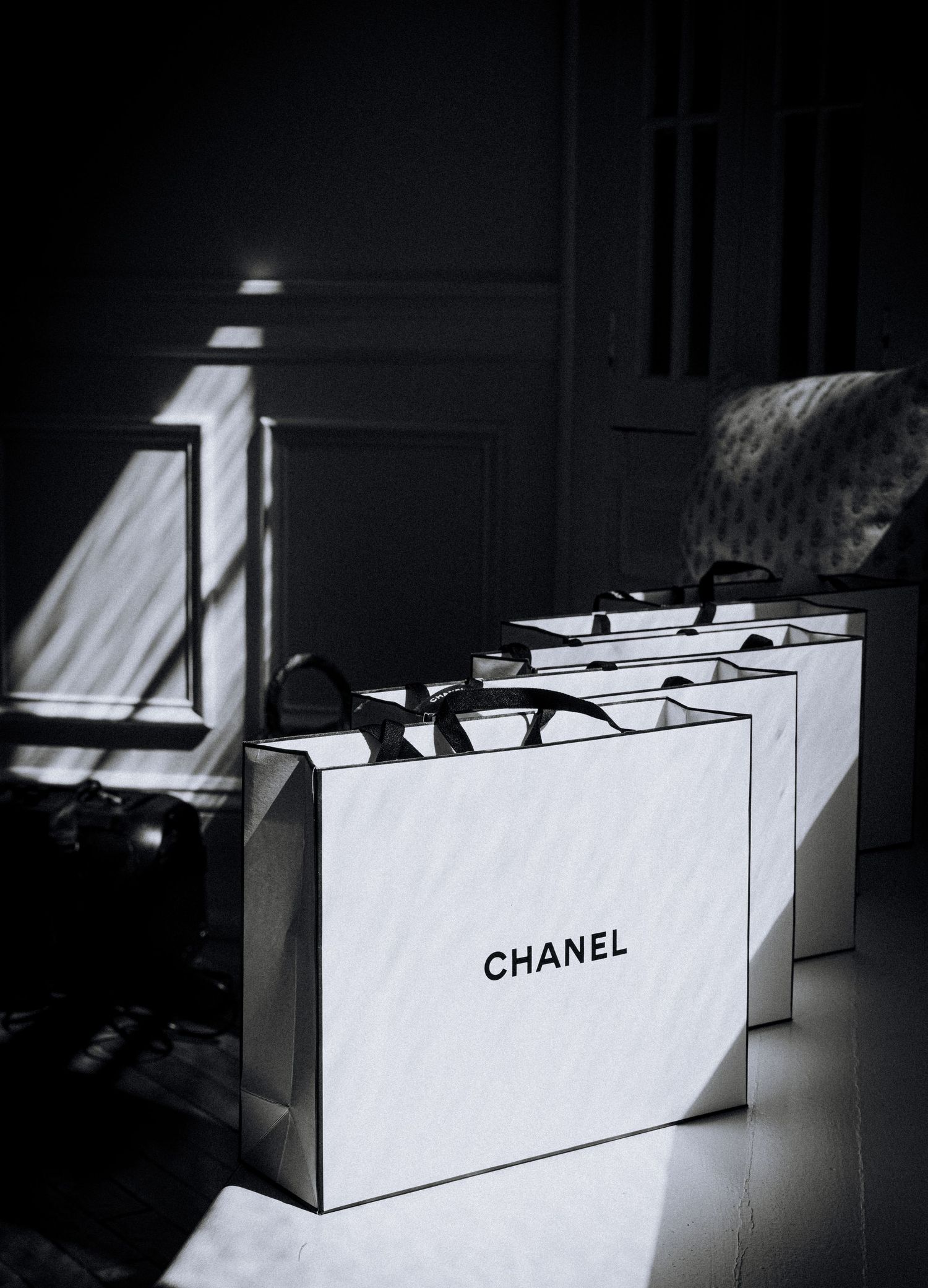 Three white Chanel shopping bags with black handles arranged in a row, captured in dramatic black and white lighting.
