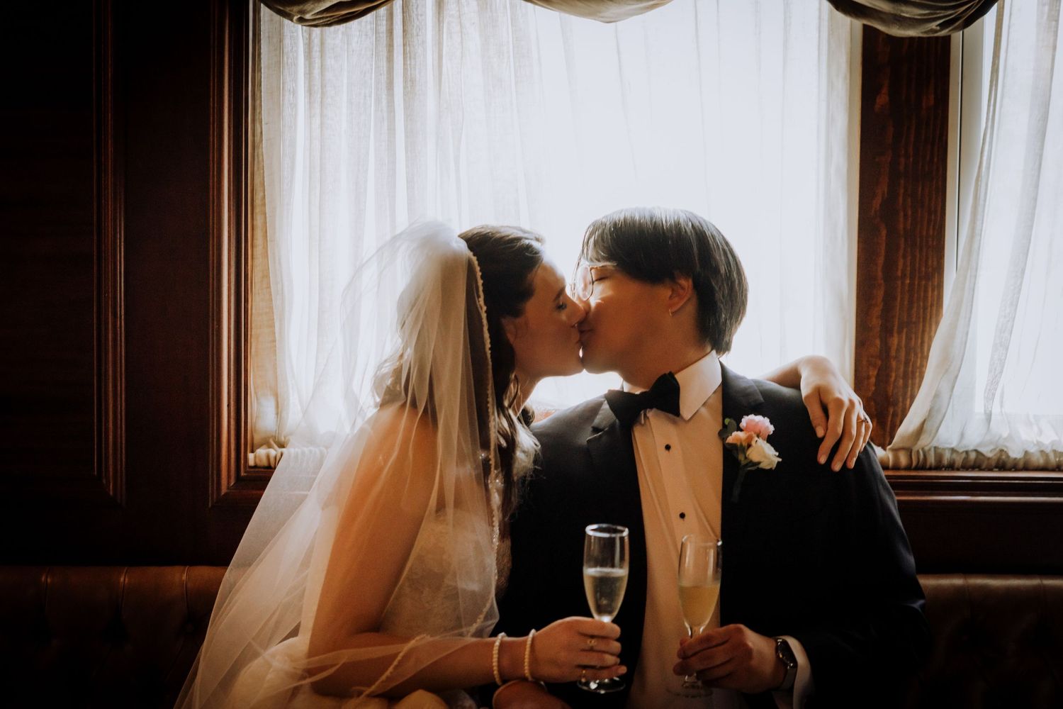 Bride in a white veil and groom in a dark tuxedo share a kiss while holding champagne flutes beside sheer curtains.