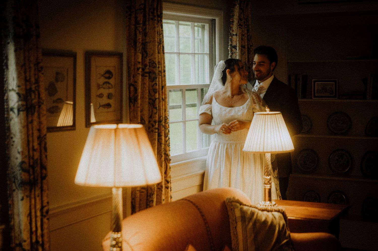 A bride and groom share a romantic moment by a sunlit window in an elegantly decorated room with warm lamp lighting.