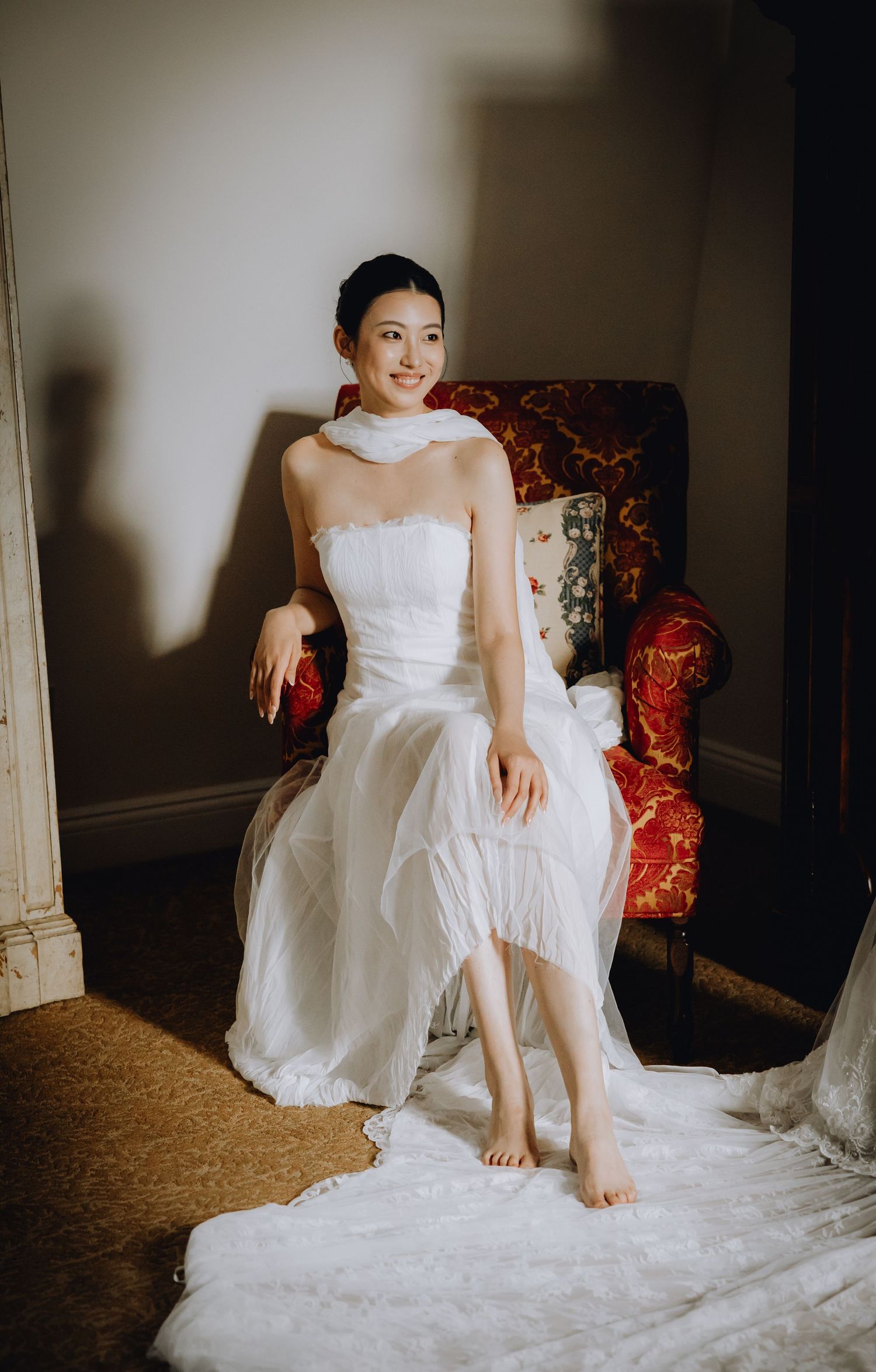 Bride in a strapless white gown with sheer scarf sits gracefully in a red floral chair in a softly lit room.