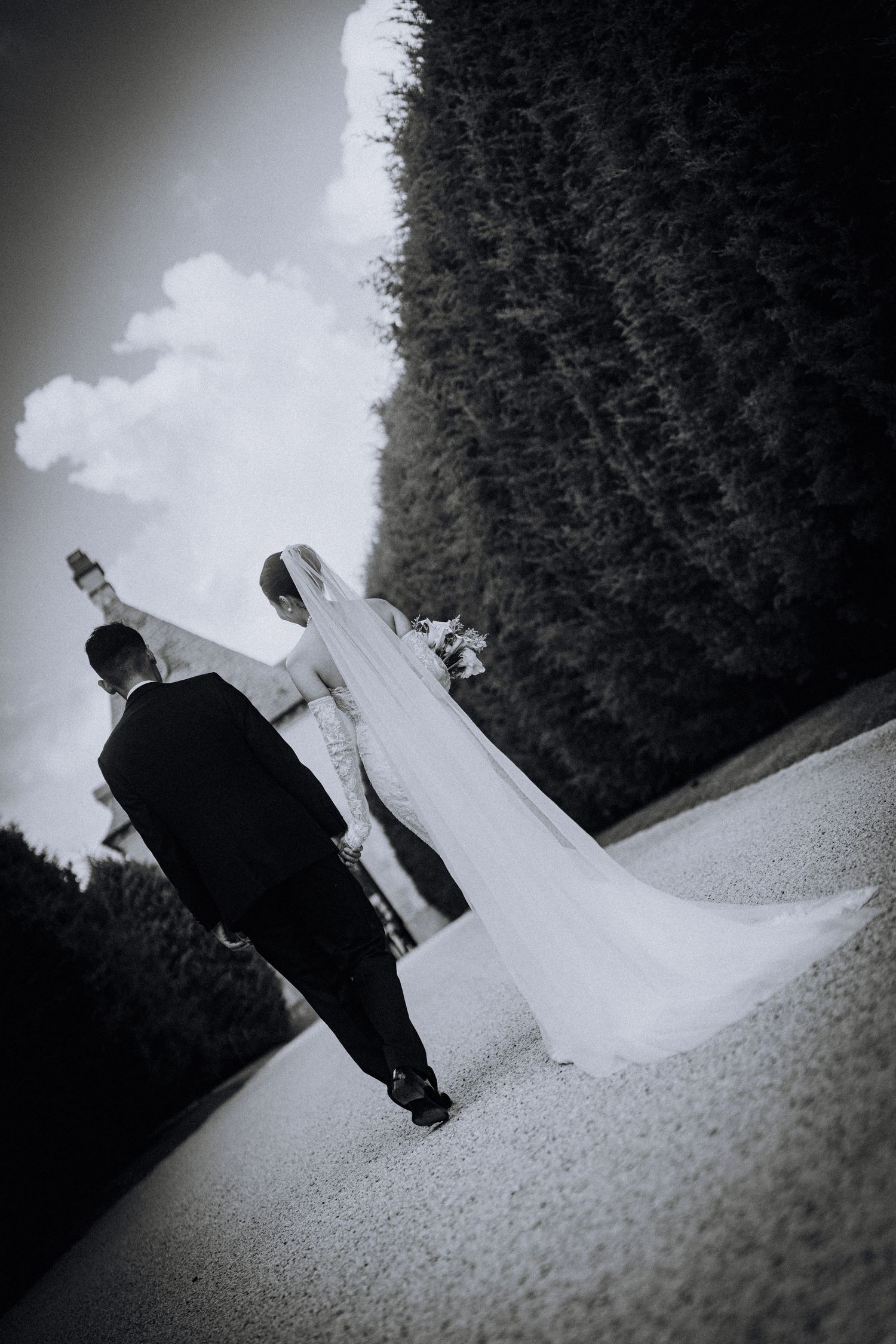 Black and white photo of a bride and groom walking away on a garden path, bride wearing a long veil and gown.