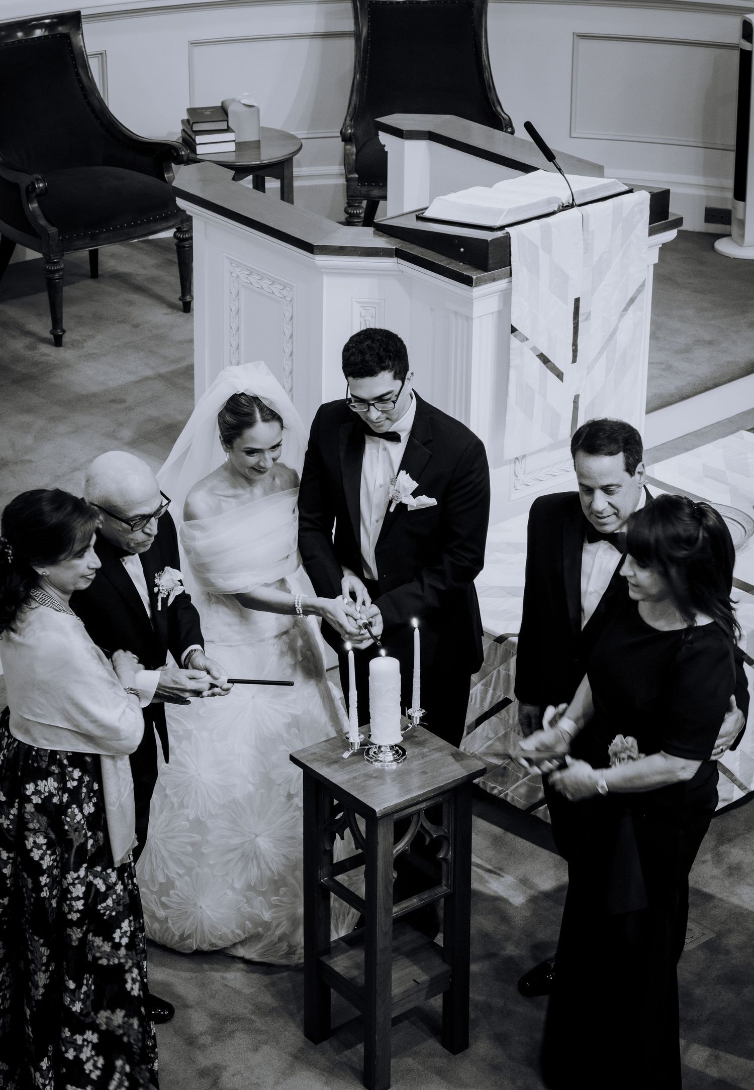 Black and white photo of bride and groom lighting unity candle at church altar surrounded by family members.