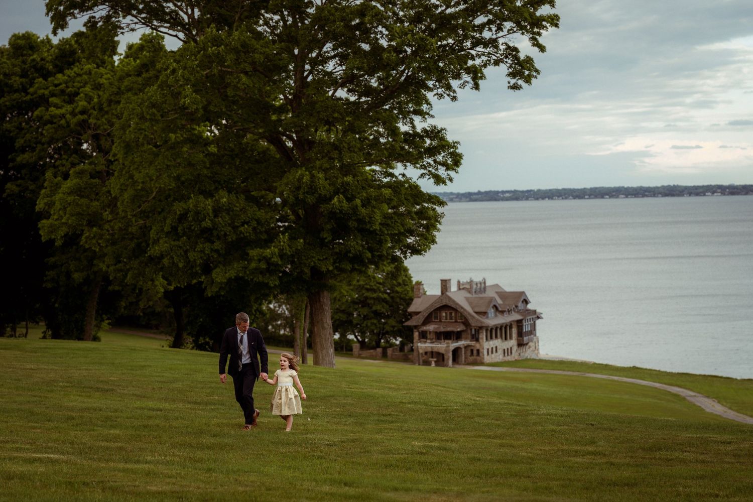 A man and child walk hand in hand on a lush green lawn beside a historic estate overlooking a calm bay.