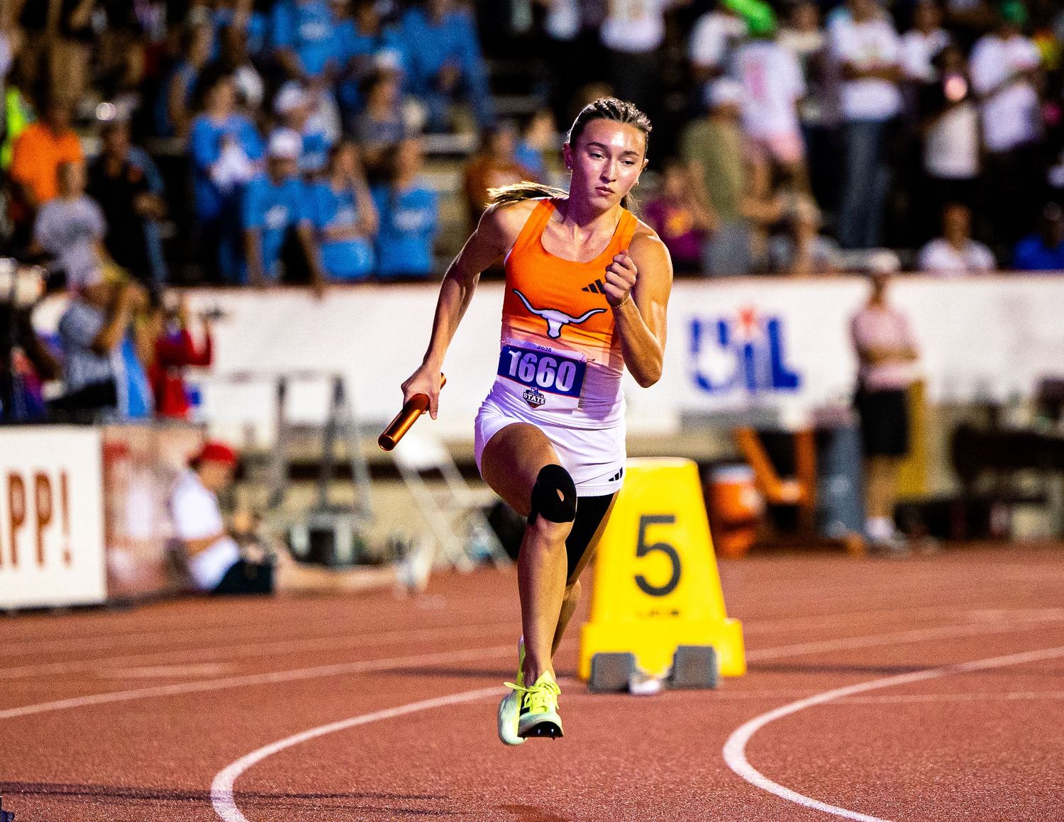 Female track athlete in orange uniform #1000 runs relay race on red track, holding baton at night UIL competition.