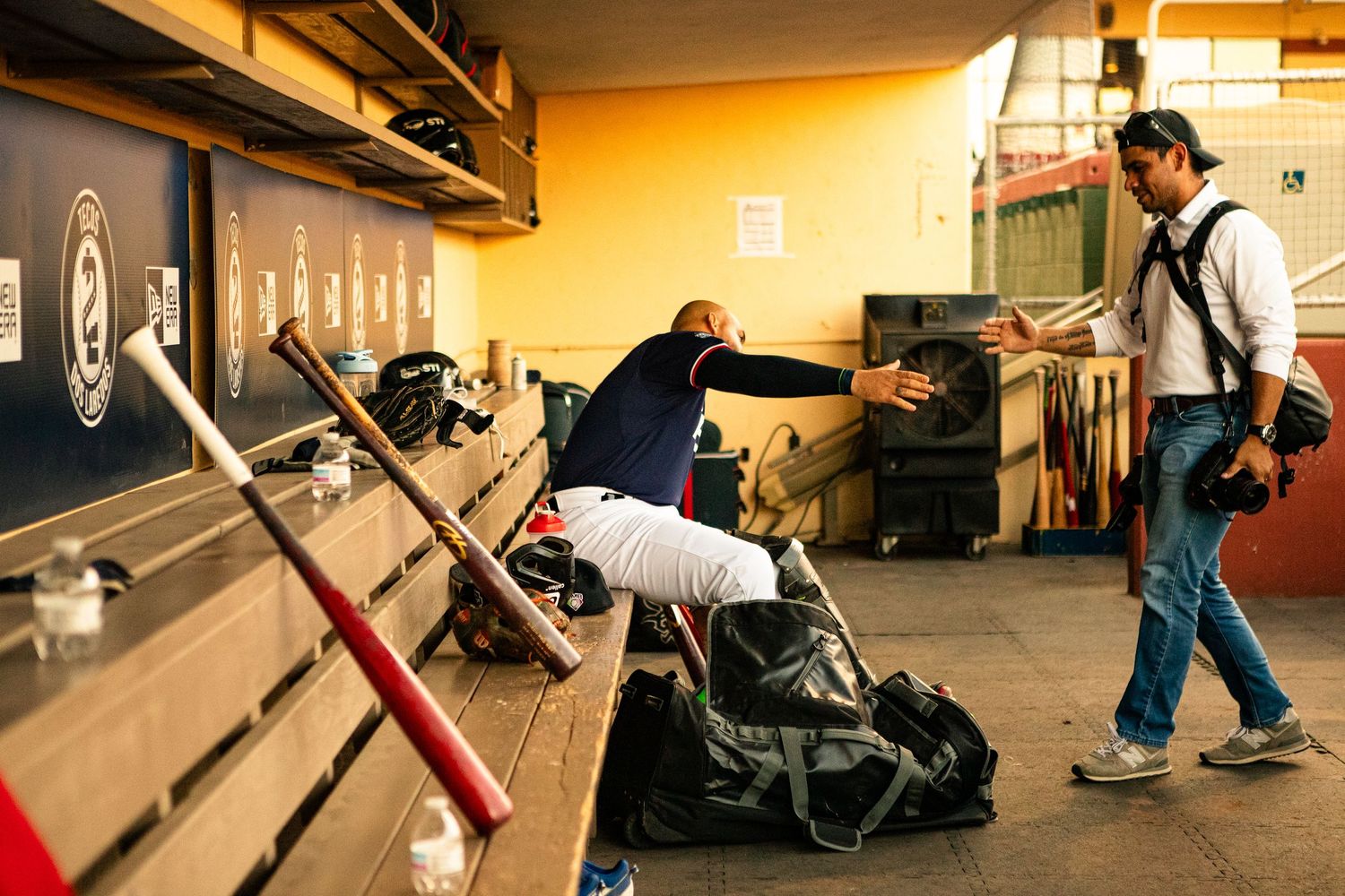Baseball player in dugout reaching out to photographer amid bats and gear in stadium locker room.