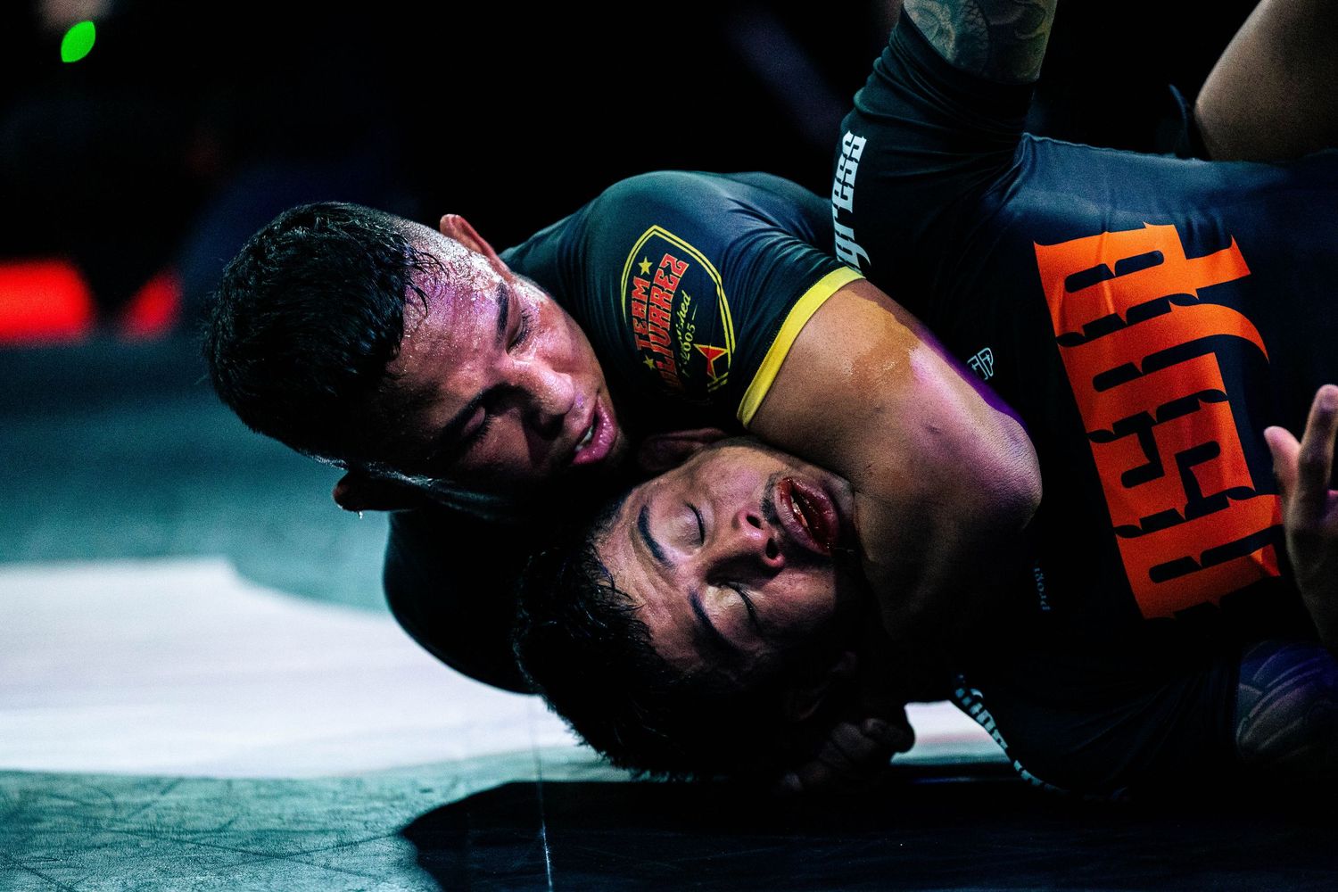Two athletes grappling in a Brazilian Jiu-Jitsu match on a competition mat with dramatic lighting.