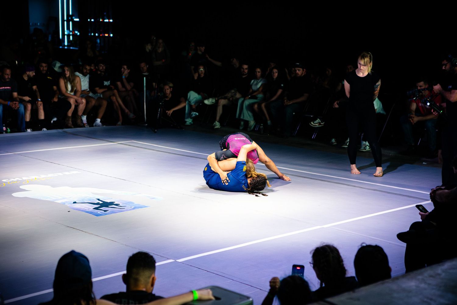 Two female athletes grappling on a lit mat during a competitive jiu-jitsu match before a live audience.