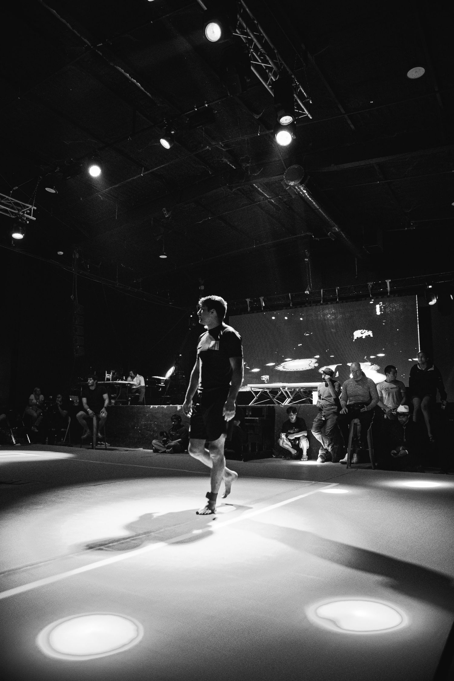 Black and white photo of a dancer performing on a lit stage with spotlights and audience in the background.