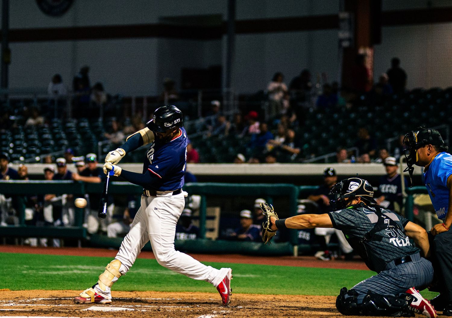 Baseball batter swings at pitch during night game, catcher crouches behind home plate in packed stadium.