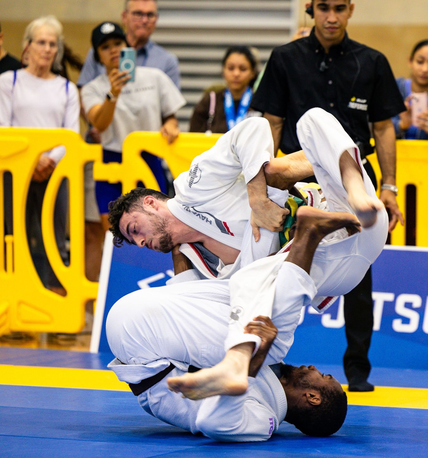 Two BJJ competitors in white gis grappling on the mat during a jiu-jitsu tournament competition.