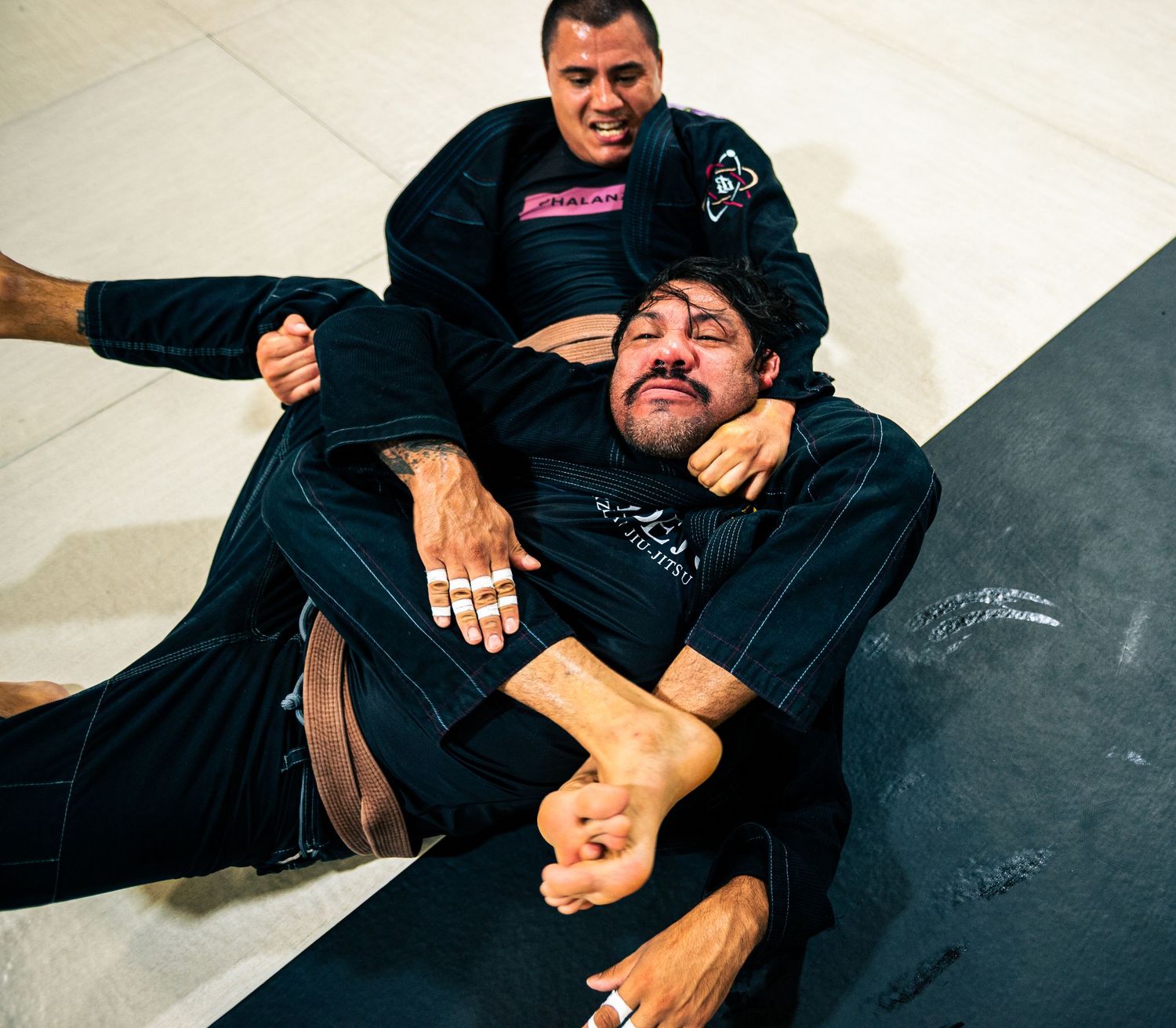 Two men in black gi uniforms practice Brazilian jiu-jitsu grappling techniques on a mat.