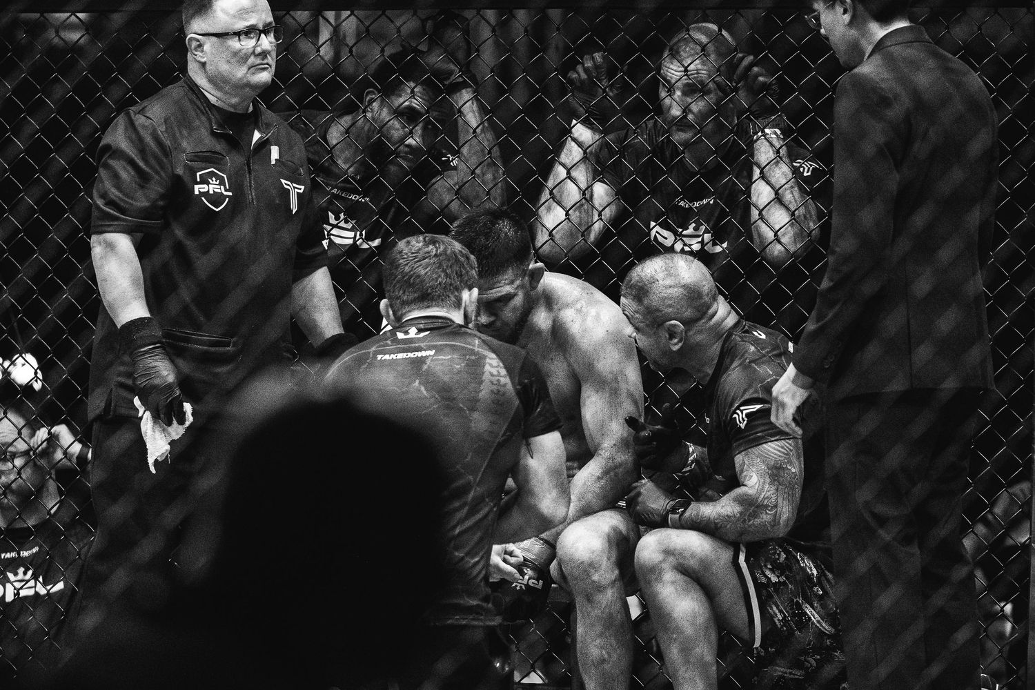 Black and white MMA fighters resting in corner of octagon cage between rounds with referee and cornermen.