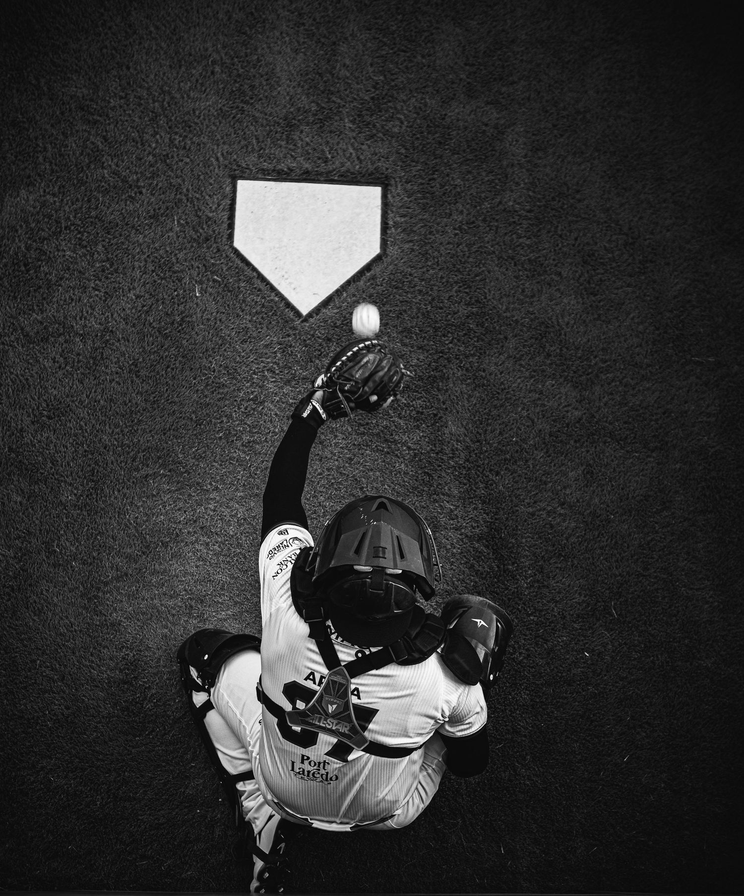 Aerial black and white photo of a baseball catcher reaching up to catch a ball near home plate.