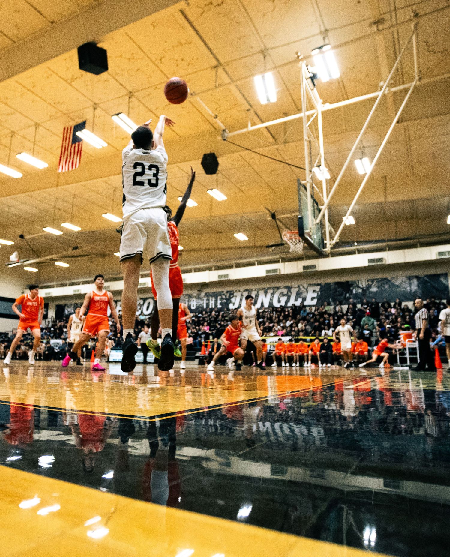 Player #23 shoots a jump shot during an indoor high school basketball game in a packed gymnasium.