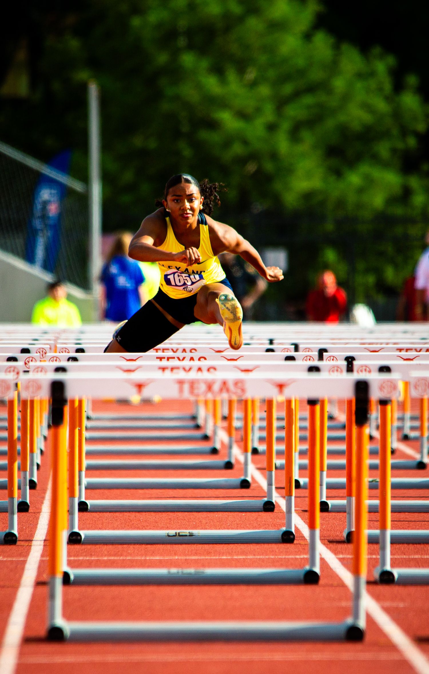 Athlete in yellow uniform leaping over a hurdle on a Texas track during a competitive hurdles race.