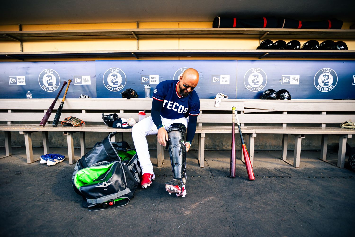 Baseball player in Tecos jersey sits in dugout preparing equipment, with bats and gear bag nearby.