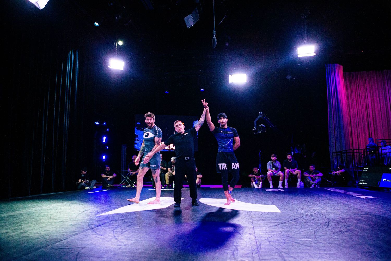 A referee raises a fighter's hand in victory on a lit stage during a martial arts competition.
