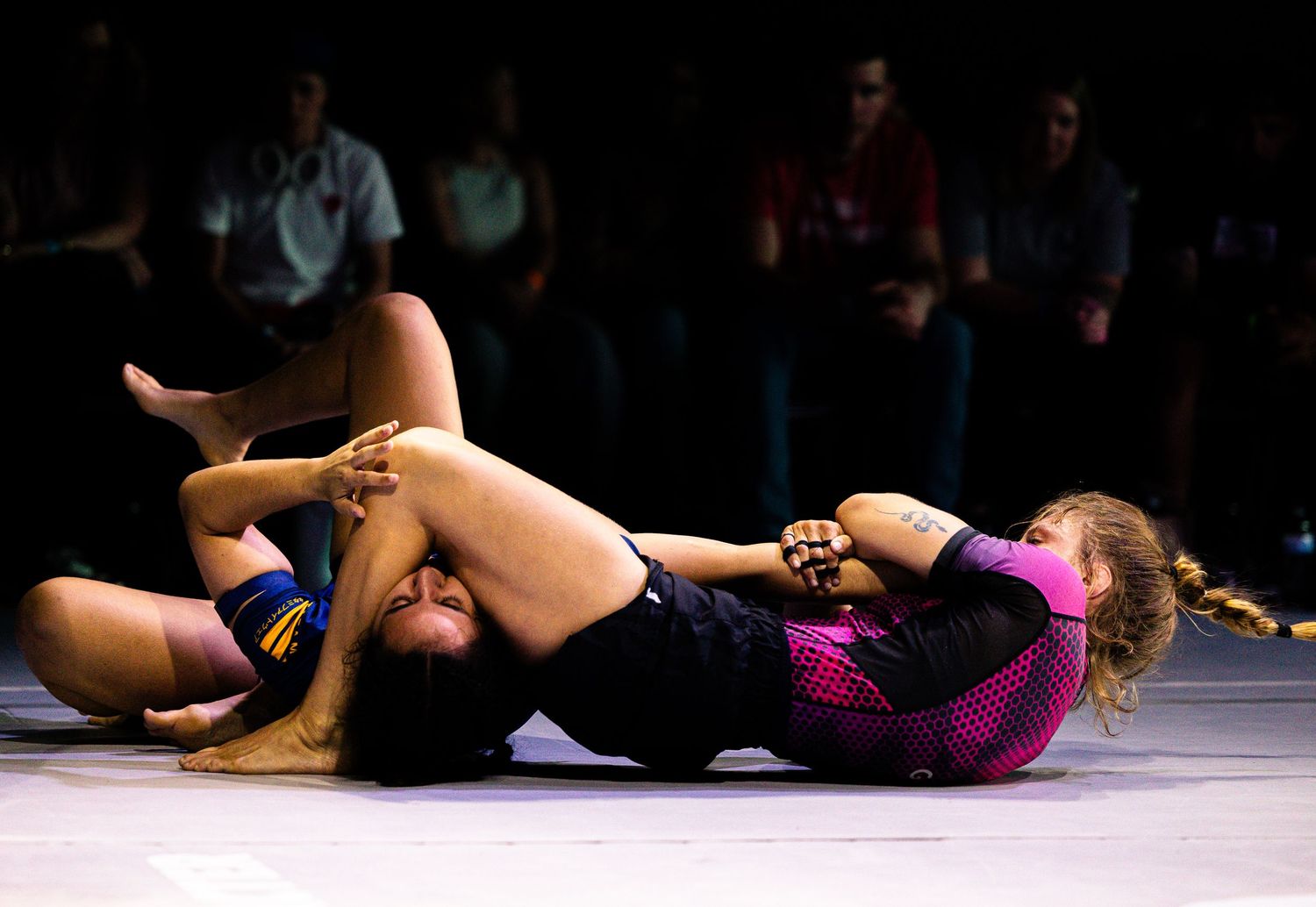 Two female grapplers compete in a submission wrestling match on the mat under bright lights.