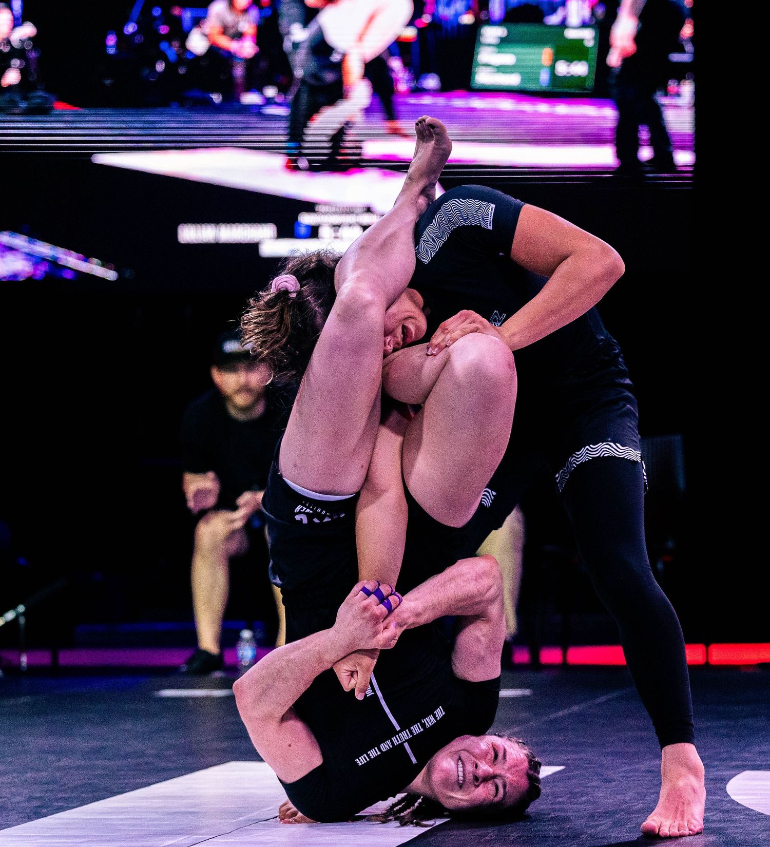 Two athletes competing in a grappling match at a professional BJJ event under bright arena lights.