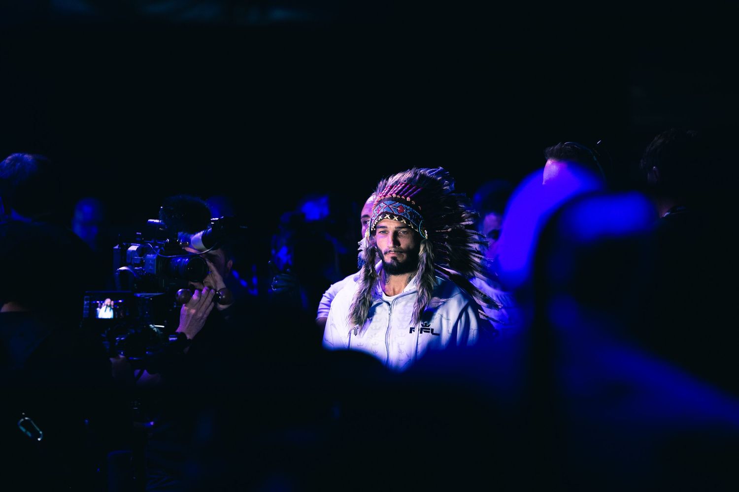 Man wearing a Native American headdress stands in a dark, blue-lit venue surrounded by a crowd.
