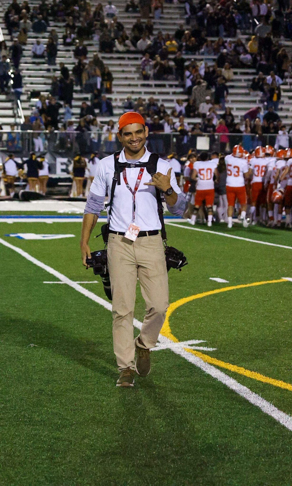 Person in khaki uniform walking along sideline of football field during game.