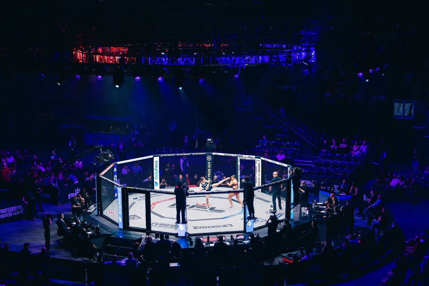 Aerial view of an MMA octagon cage illuminated by bright lights in a packed arena during a fight event.