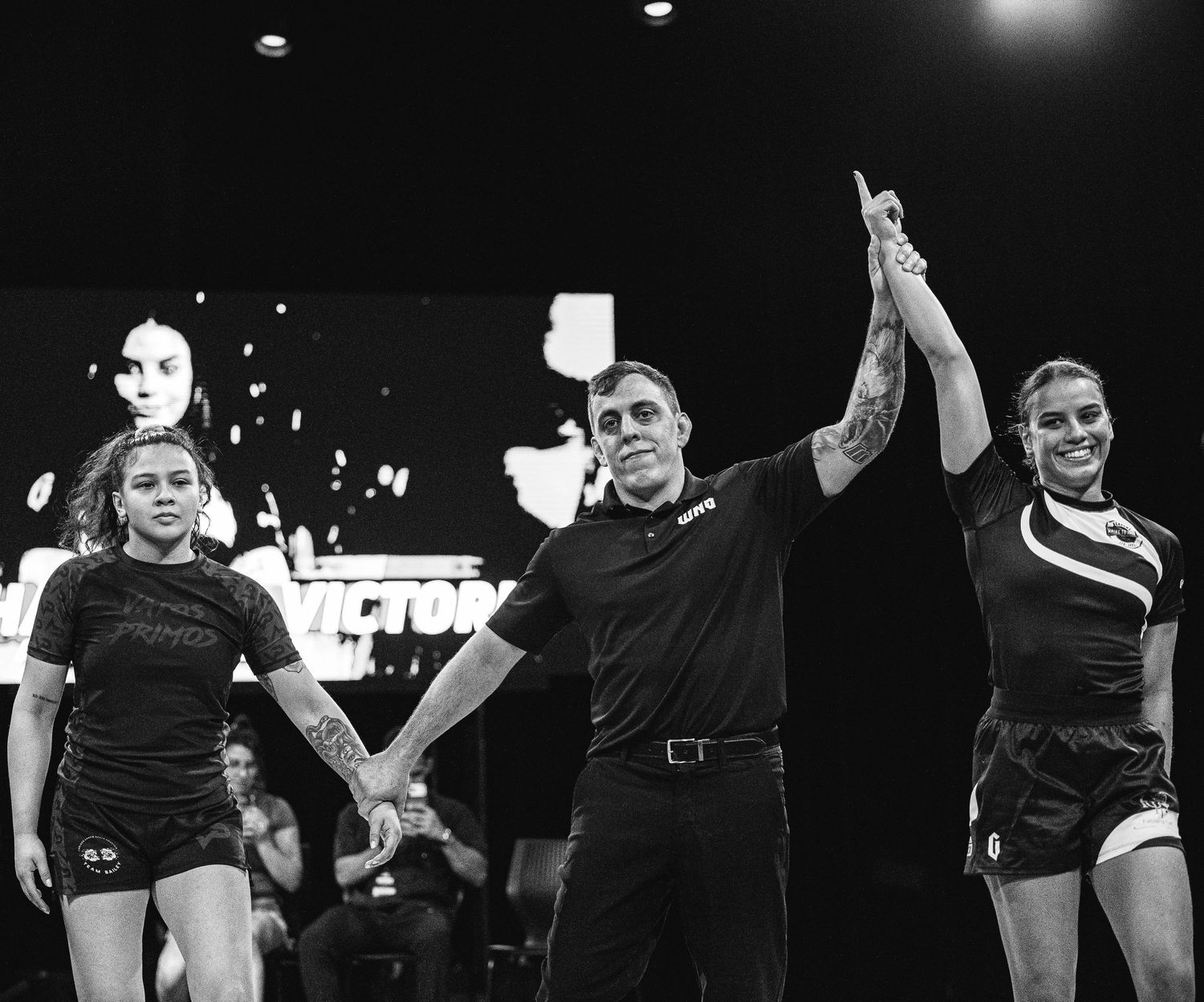Referee raises winner's hand at grappling tournament as two female competitors stand on stage under bright lights.