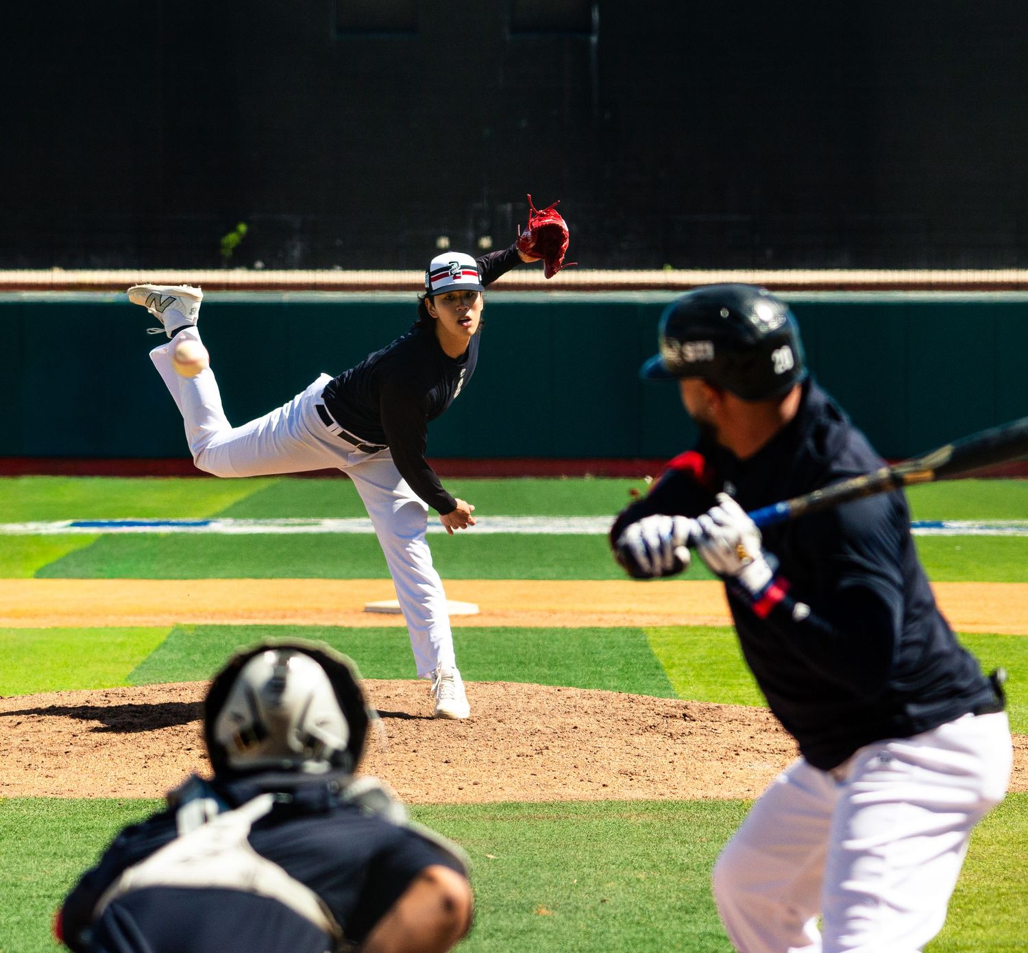Baseball pitcher mid-throw on mound with batter ready at plate and catcher crouching during sunny outdoor game.