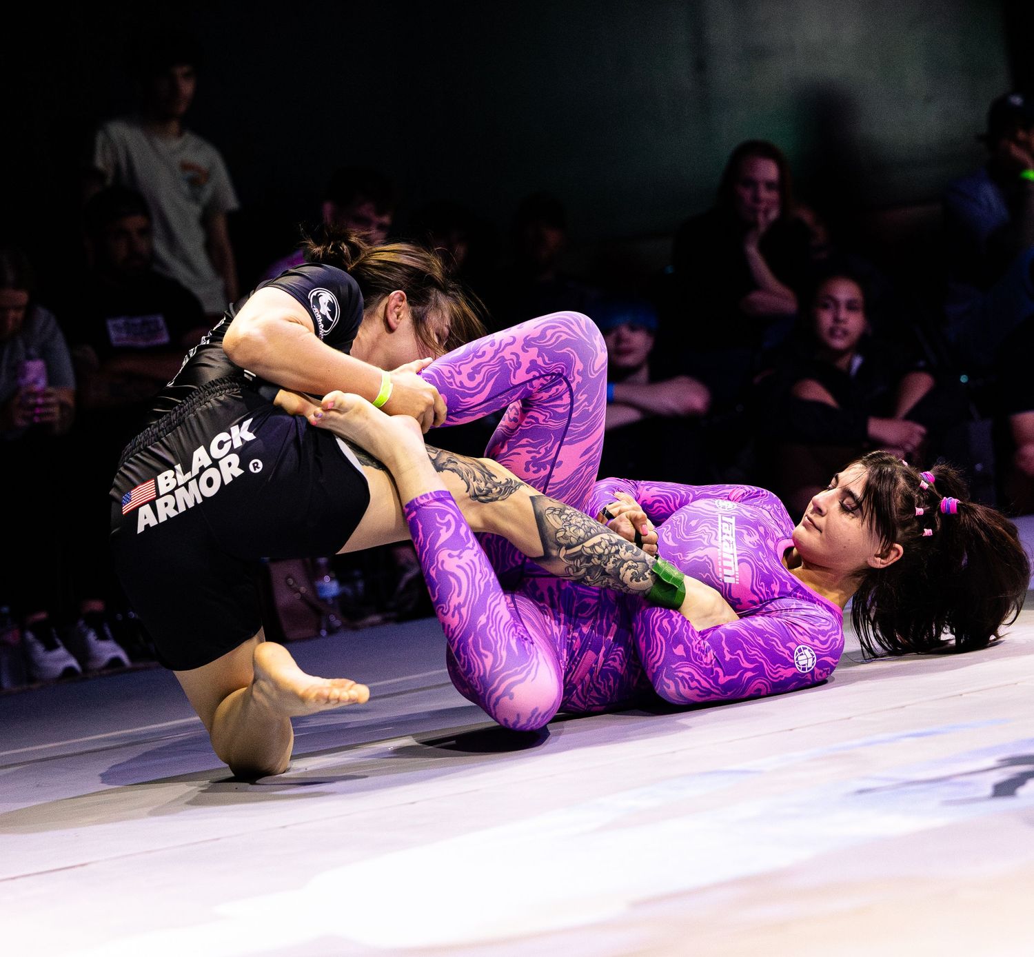 Two women compete in a grappling match, one in purple spandex and one in black, on a white mat at an MMA event.