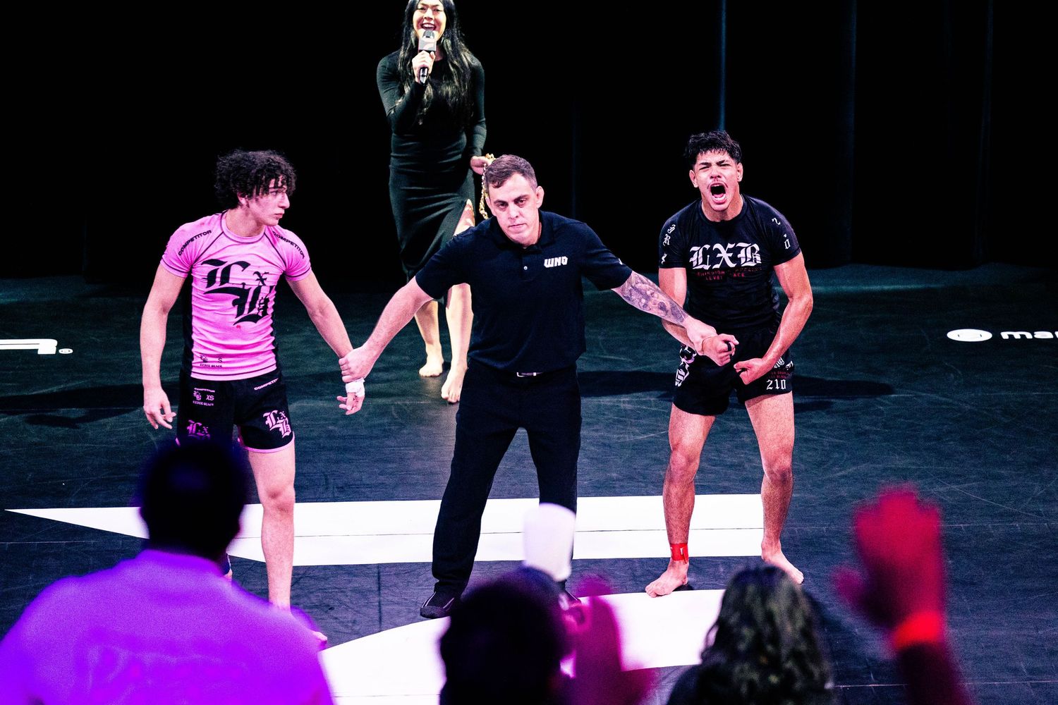 Referee raises hands of two fighters in pink rash guards at an MMA grappling event on a lit stage.
