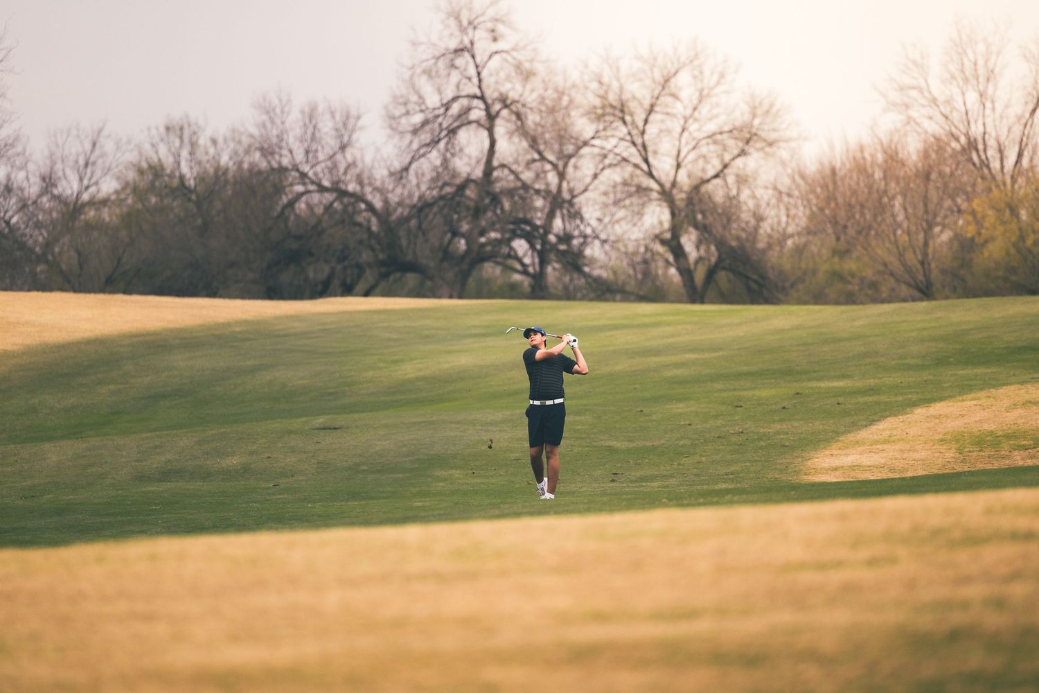 Golfer mid-swing on a lush green golf course fairway surrounded by trees on a sunny day.