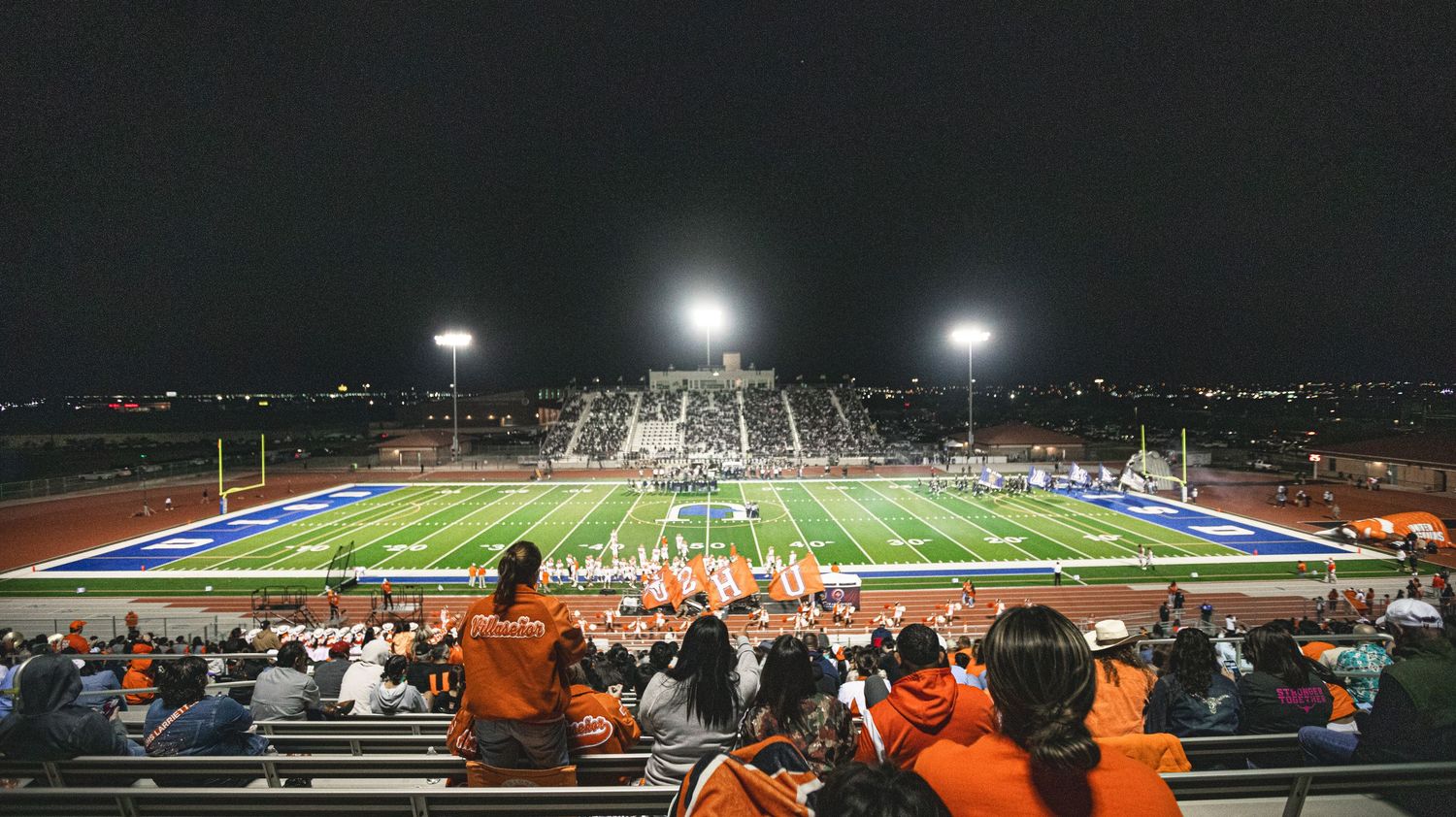 Night football game at a high school stadium with fans in orange cheering under bright lights on a blue turf field.
