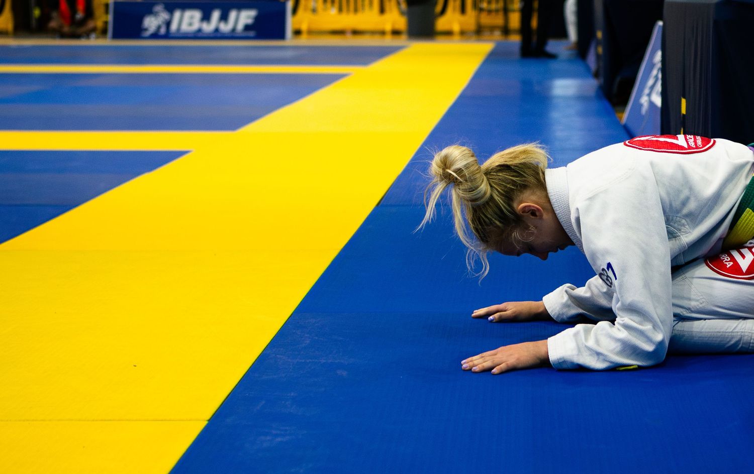 Female BJJ competitor bowing on blue and yellow IBJJF competition mat during jiu-jitsu tournament.