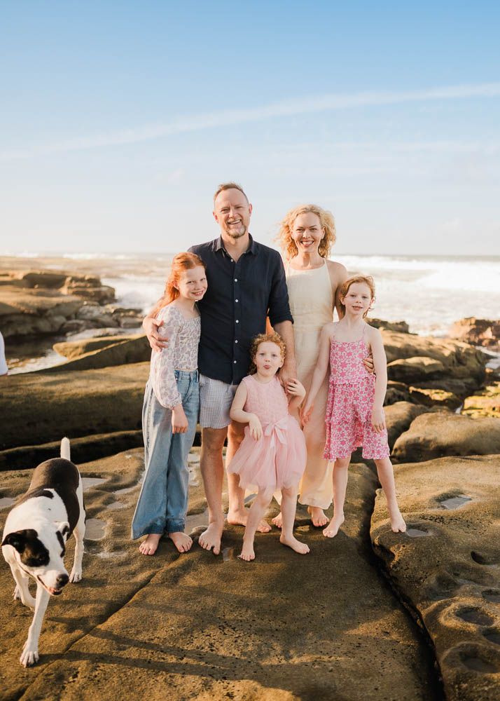 Family of five with a dog posing on rocky beach shore with ocean waves in the background on a sunny day.