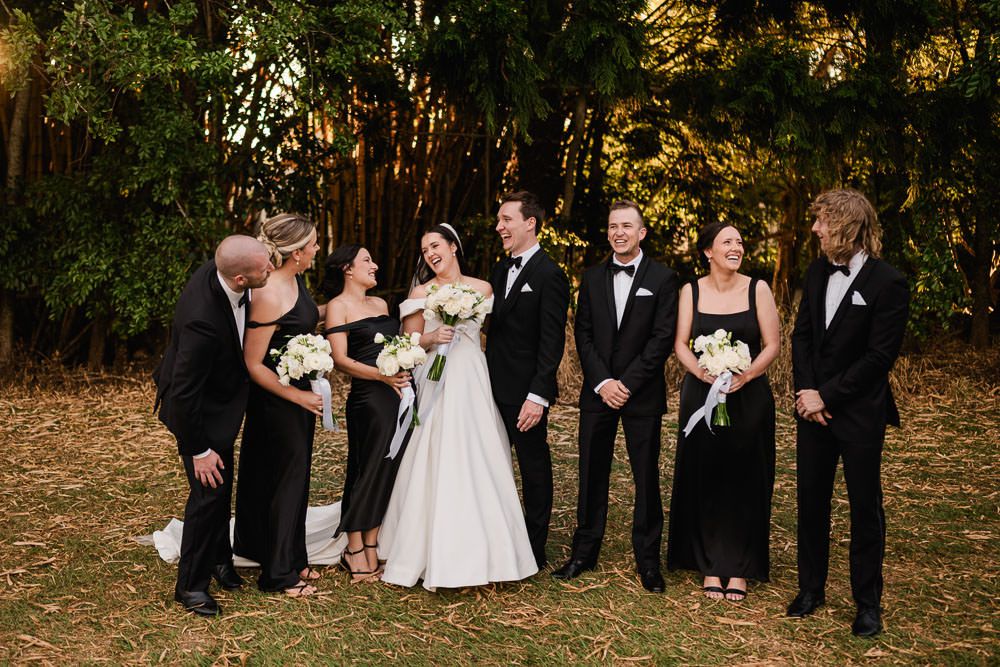 Wedding party in formal black and white attire poses together on autumn day with fallen leaves in background.