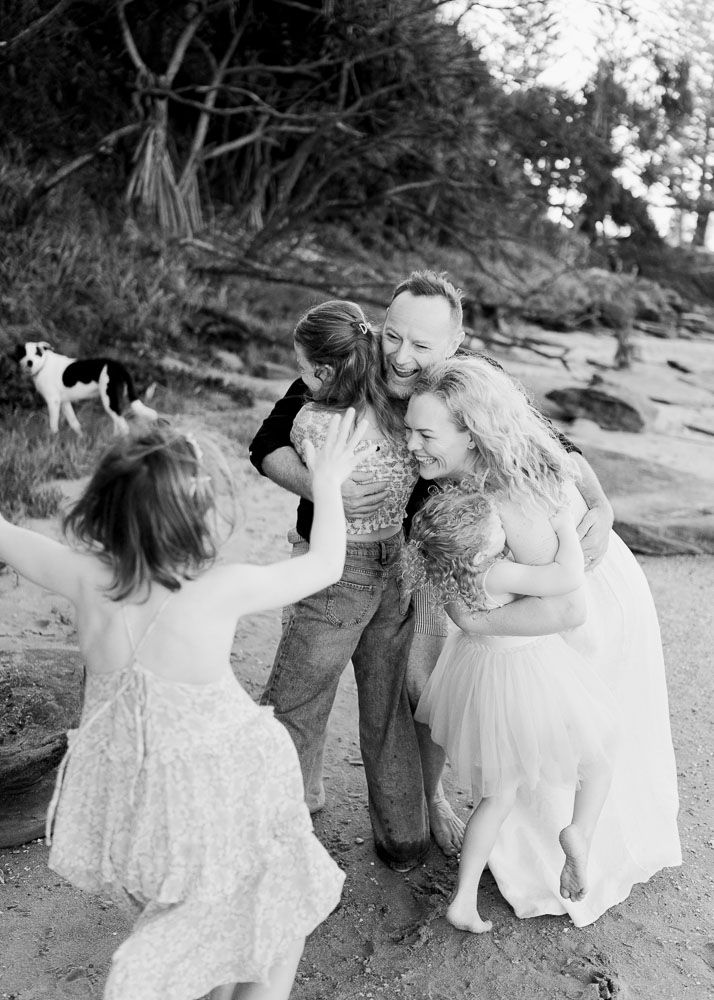 Black and white photo of a joyful family embracing on a sandy beach with trees and a dog in the background.