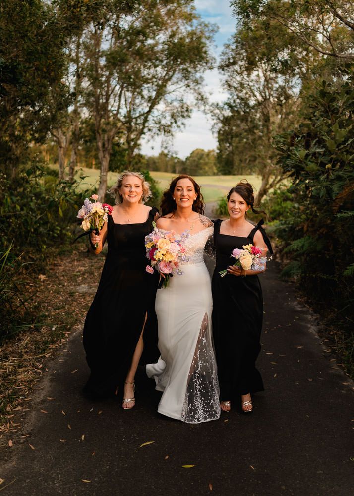 Three women in formal attire walking and laughing together on a tree-lined path while holding colorful bouquets.