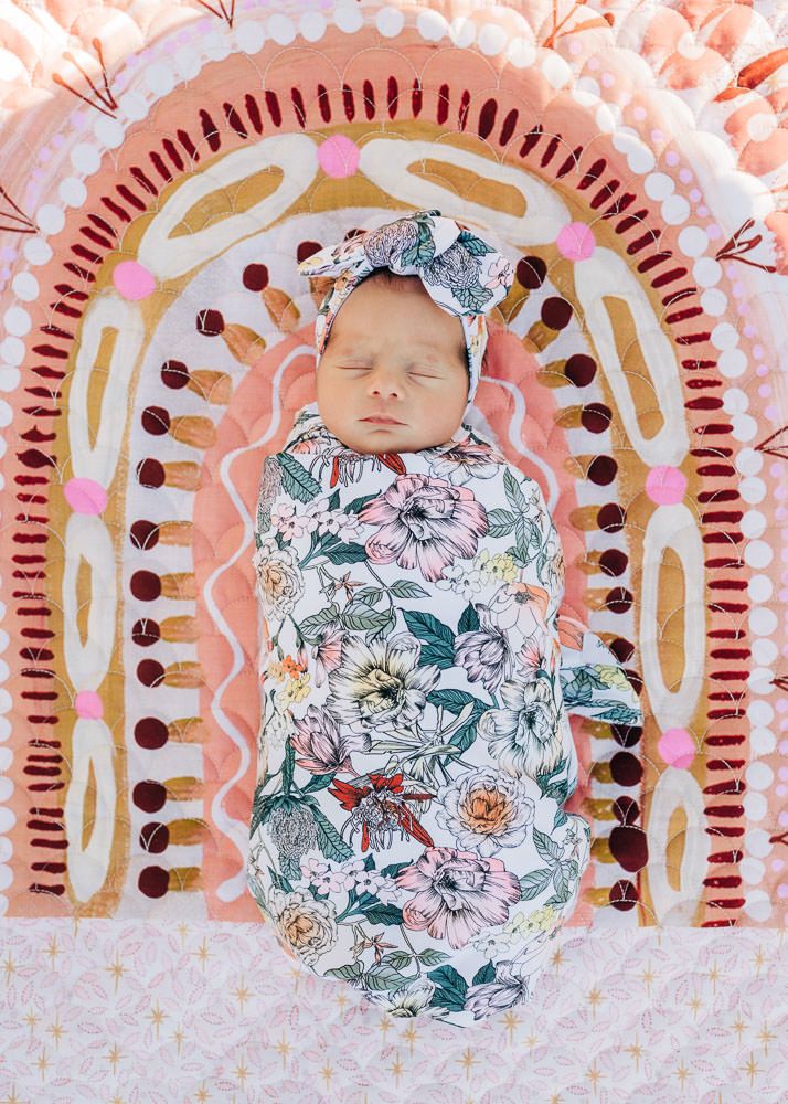 A sleeping baby swaddled in floral print fabric lies on a pink background with decorative boho rainbow arch design.