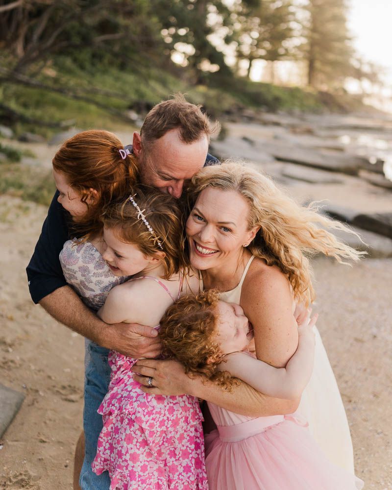 Happy family of five sharing a warm group hug on a sandy beach at golden hour.