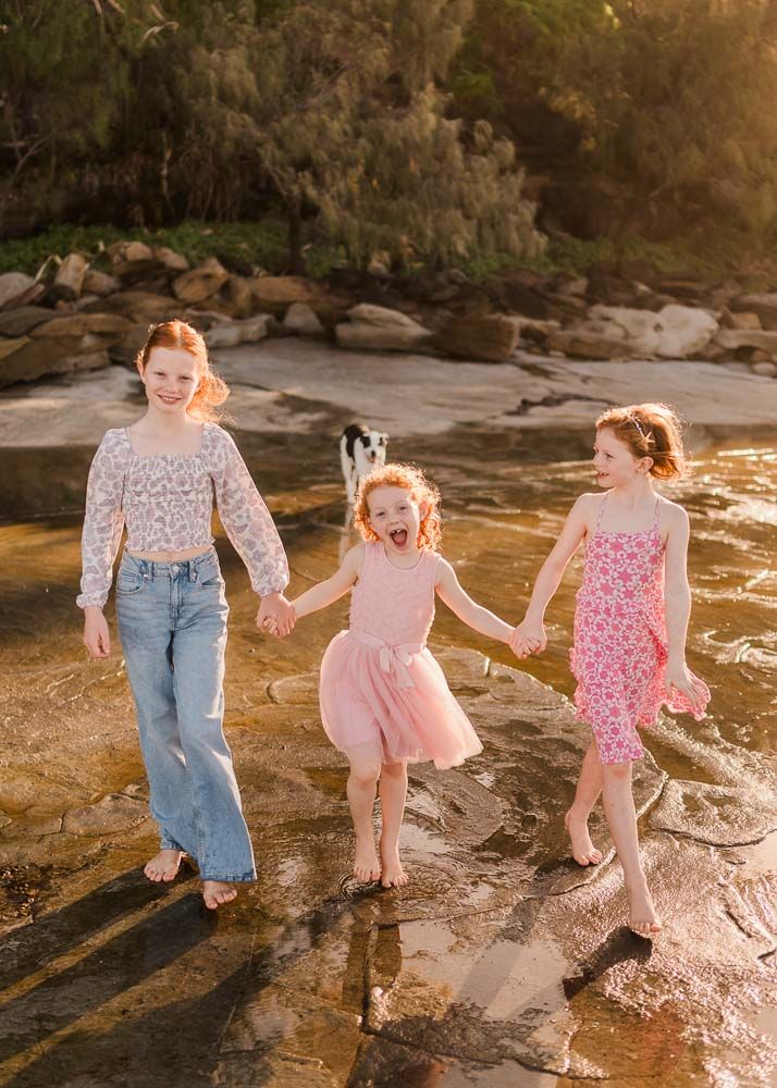 Three happy girls holding hands and walking barefoot on wet rocks near a beach with golden sunlight and trees behind them.