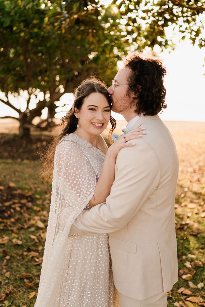 A couple embraces outdoors at sunset, wearing light colored formal attire under a tree with golden light filtering through.
