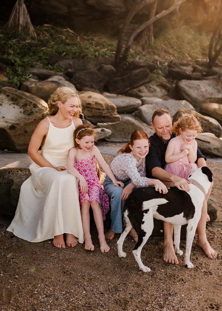 Happy family with three girls and a black and white dog sitting together on rocks at a beach.