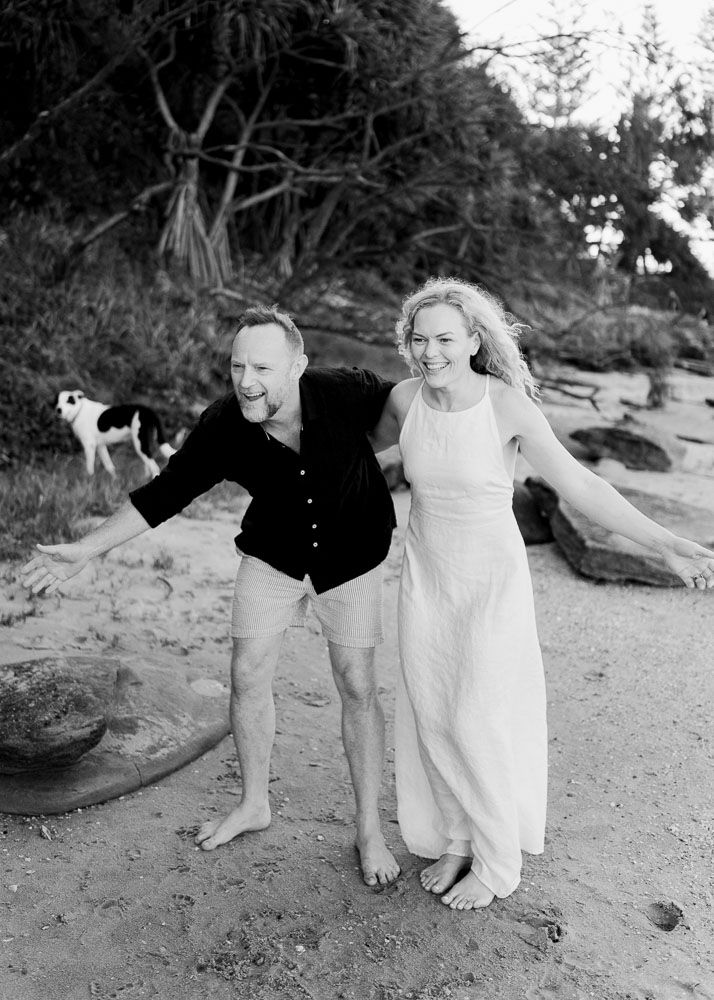 Black and white photo of a laughing couple in white dress and black shirt walking barefoot on a sandy beach.