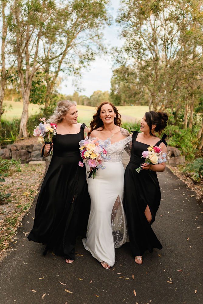 Three women in black and white dresses share joyful moments while walking outdoors with floral bouquets.