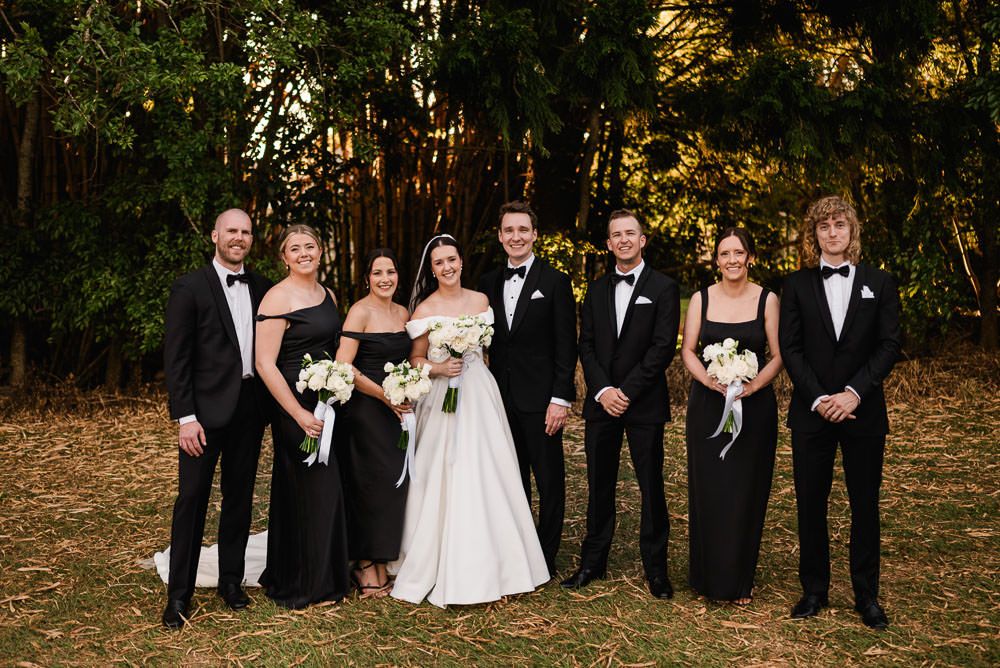 A wedding party in formal black and white attire poses together outdoors during autumn.