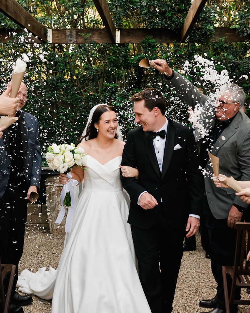 A joyful wedding celebration with confetti being tossed as a couple walks down an outdoor garden path together.
